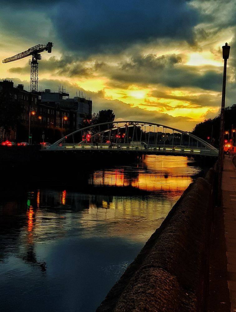 Sunset over river Liffey. 
River wall on the right. A bridge crossing the river. Terrific clouds in grey in the sky. Orange and yellow light in the sky reflect in the river. 