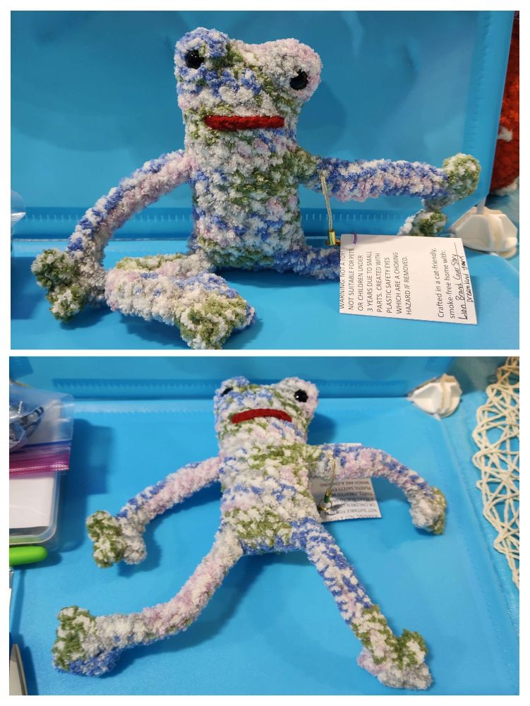 Two photos of a crocheted "leggy" frog, which is a frog with a longer body and longer, curly legs. In the upper photo, the frog is sitting with one leg bent and one leg out, whereas in the bottom photo, the frog is laying on its back. The frog itself is made out of a mixed green, blue, and white blanket yarn, and has two black beady eyes at the top of its head. It has a wide, deep red strip of red yarn on its face that resembles a mouth. 
In both photos, the frog is in front of a light blue backdrop.
