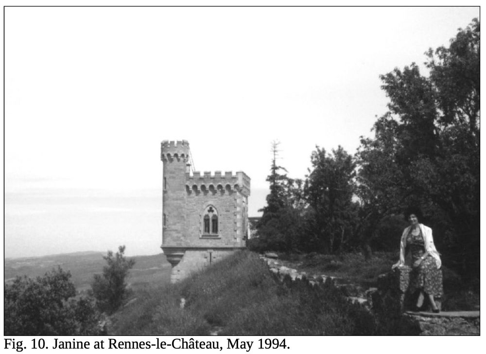Black and white photo focusing on a square medieval tower attached to a cliff, trees surrounding the landscape on the right as the cliff falls away to the left and a vast horizon behind it. A woman in a dress and white sweater draped over her shoulders sits on a stone wall in the foreground right. Caption: "Fig. 10. Janine at Rennes-le-Château, May 1994."
