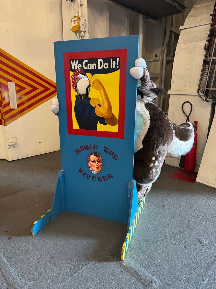A crocodile-deer hybrid fursuit poses with their snout poking out of the facehole of one of those cutout standees representing Rosie the Riveter, the lady doing an arm pump while saying “We can do it!”