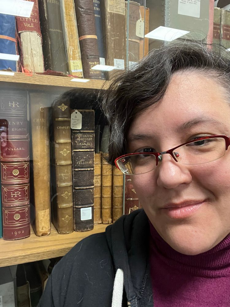 Valentine X smiling while posing in front of a bookshelf filled with antique and historical books, including titles about Henry VIII and Charles I, enclosed in a glass case. The books have leather and ornate bindings.