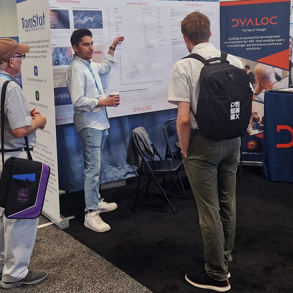 Josh David presenting APL concepts at the Dyalog booth during JSM 2025, speaking to attendees in front of the TamStat and symbolic notation posters.