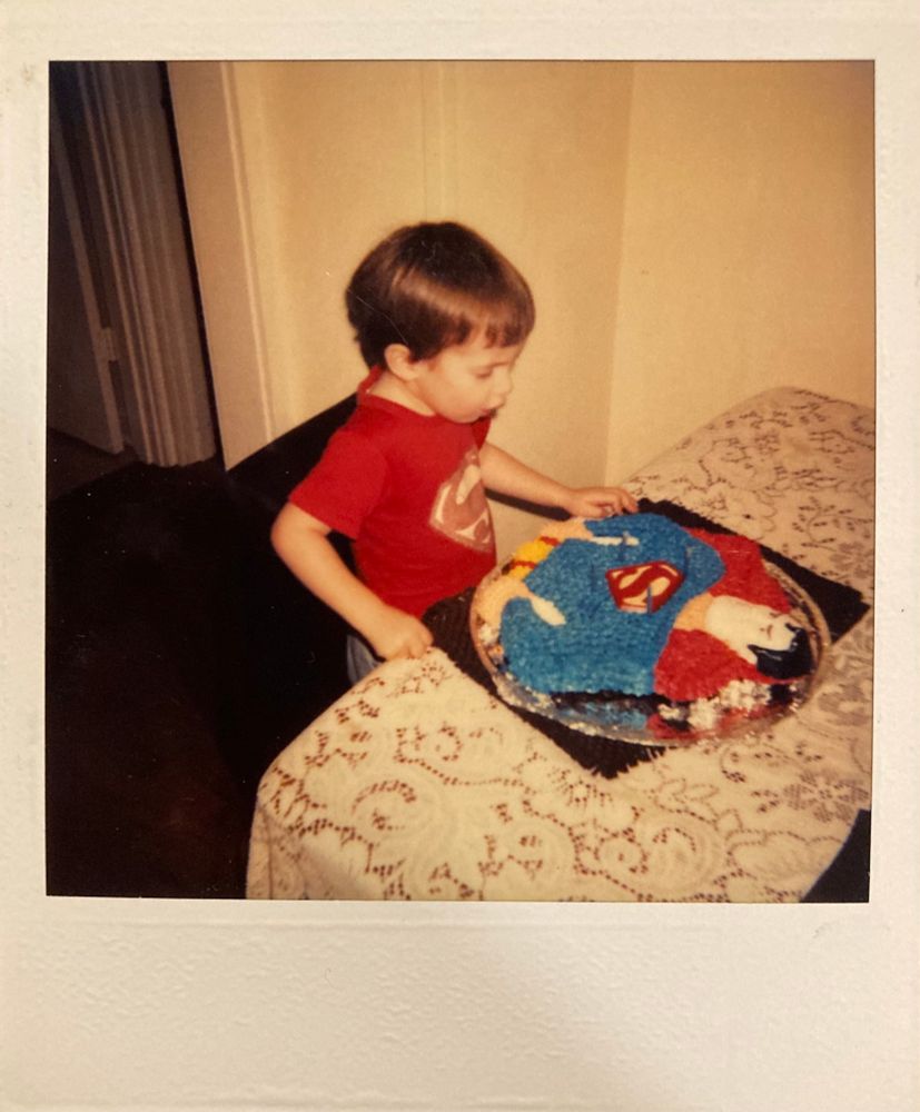 me as a little kid wearing a Superman shirt and staring down at my Superman birthday cake.
