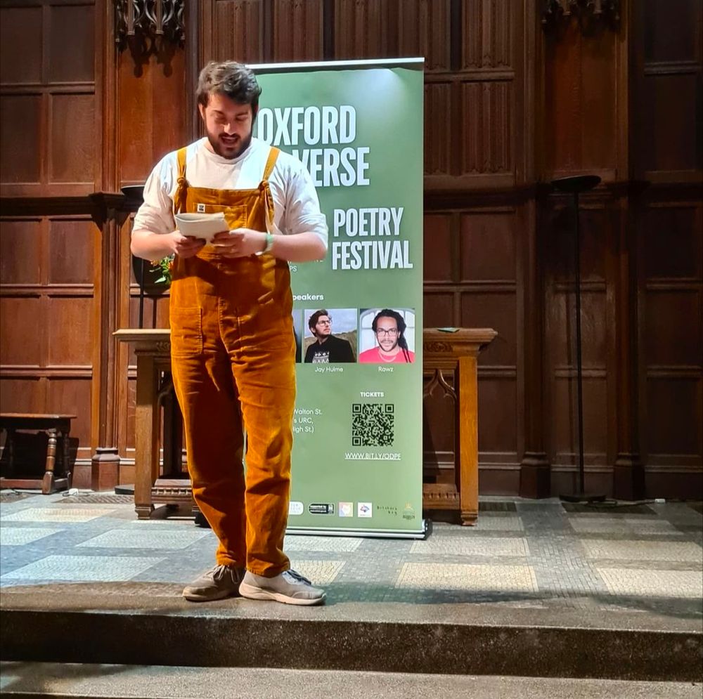 Josh standing on a platform in front of a sign that says 'Oxford Di-Verse Poetry Festival' reading from a booklet.