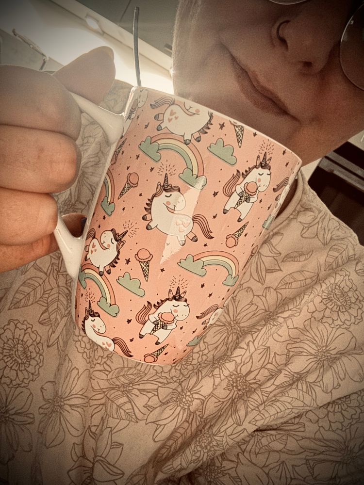 Ich halte eine große, rosa Tasse mit niedlichem Einhorn-Muster in der Hand. Die Einhörner sind mit Regenbögen, Wolken und Eistüten verziert. Im Hintergrund ist ein florales Muster auf der Kleidung erkennbar. 