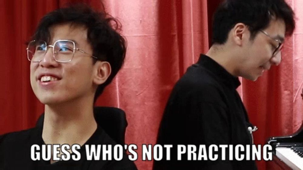 a man with glasses is smiling next to another man who is playing a piano and the caption guess who 's not practicing