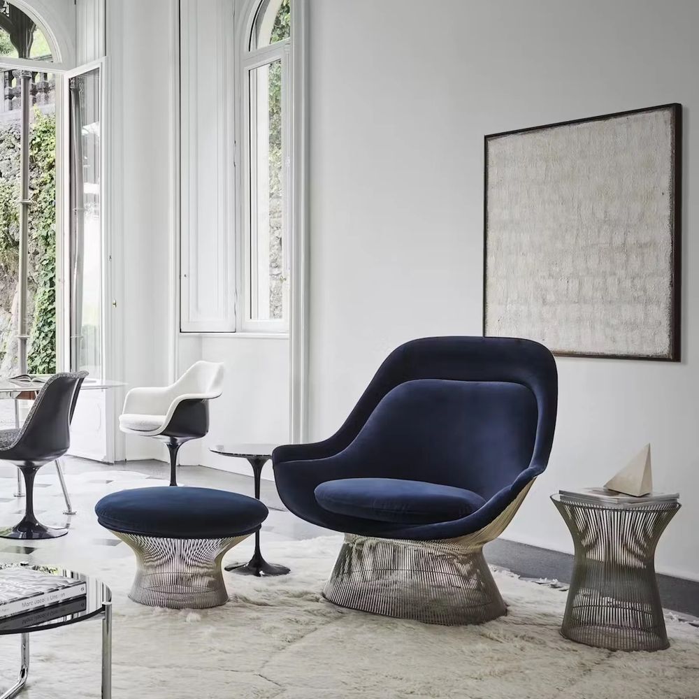 Staged shot of one of Warren Platner’s armchair designs, along with a matching ottoman and side table. They’re all based on frames made of silver metal wires connecting circular bases to circular tops, kind of hourglass-like. The armchair and the ottoman have navy blue velvet cushions. 

There are some tulip tables and chairs in the background, and large windows onto a garden
