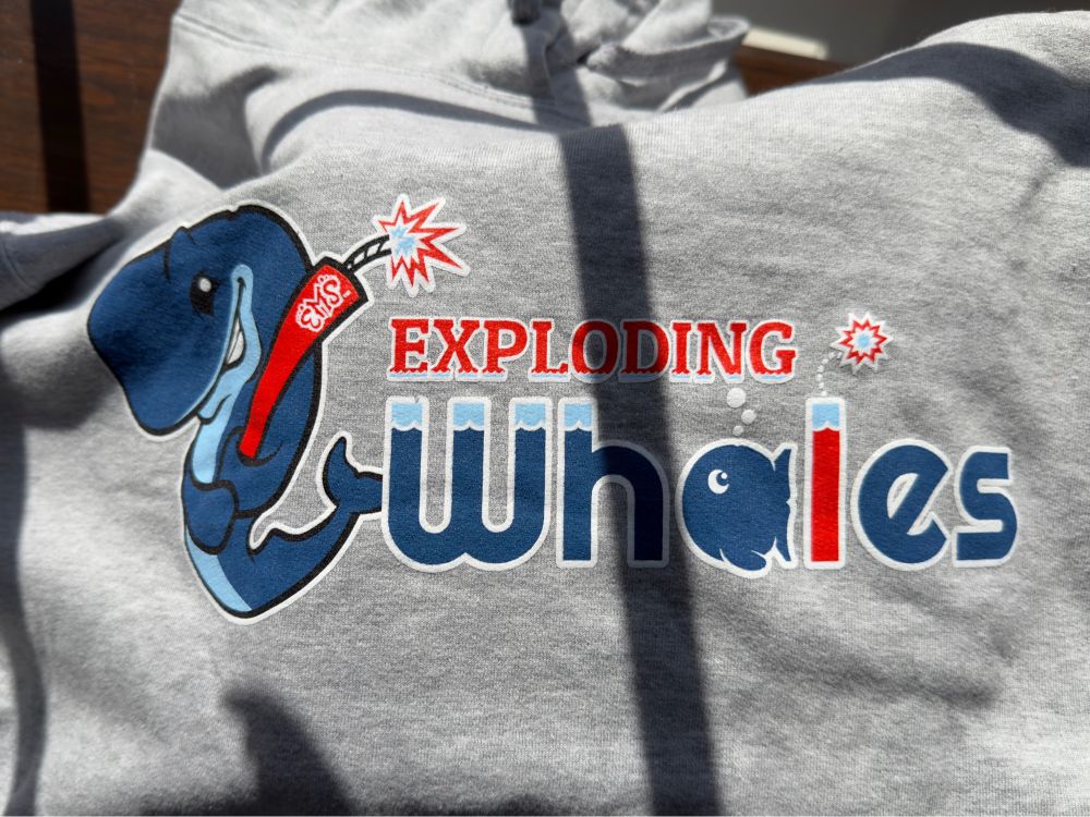 A grey hoodie from the Eugene Exploding Whales, a retheme of their minor league baseball team. It has a blue sperm whale holding a lit stick of dynamite like a baseball bat and the text “Exploding Whales” where the a is a whale silhouette and the l is another lit stick of dynamite