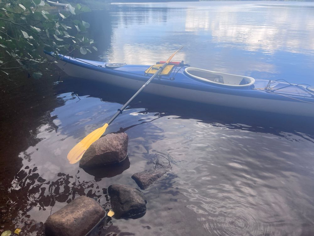 Kajakki kellumassa vedessä, mela nojaamassa kajakkiin. Pilvet heijastuu vedestä kajakin takana. Edessä kiviä ja lehdet roikkuu puusta kuvan vasemmassa ylälaidassa.