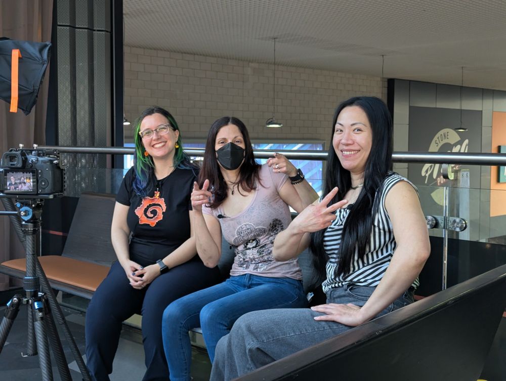 Picture of Marylia Gutierrez with green hair, a black shirt with an orange Grafana logo G, Adriana Villela with a black face mask and pink shirt, and Reese Lee with a black and white striped shirt. They are sitting on a couch over a walkway with camera gear setup for recording.