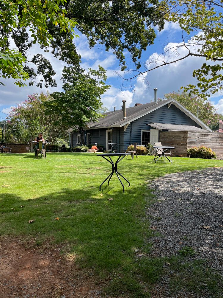 View of a local coffee shop from the backyard. Small tables are scattered across the yard with a couple of people seated at some. It’s partly cloudy and sunny outside. 