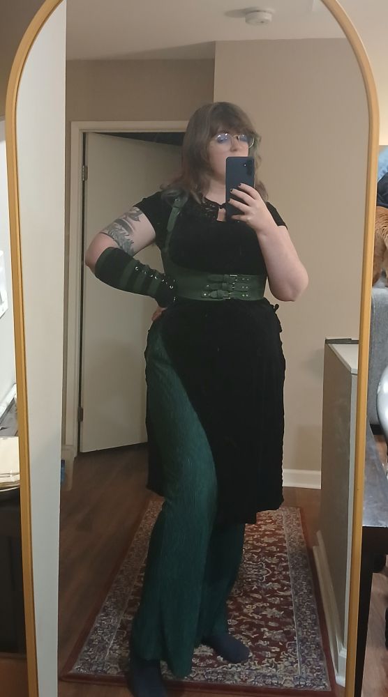A person takes a mirror selfie in a long mirror with a gold trim. She is wearing a black, short velvet dress with a slit to the hip and green trousers. Around their waist is a green corset belt and on one arm is a green and black striped arm covwr.