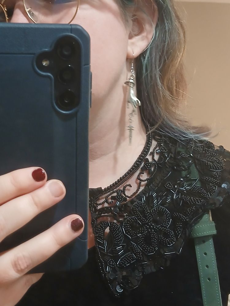 A close up mirror selfie featuring a black collar made of lace and beadwork, and an earring made of a silver hand that dangles a sword