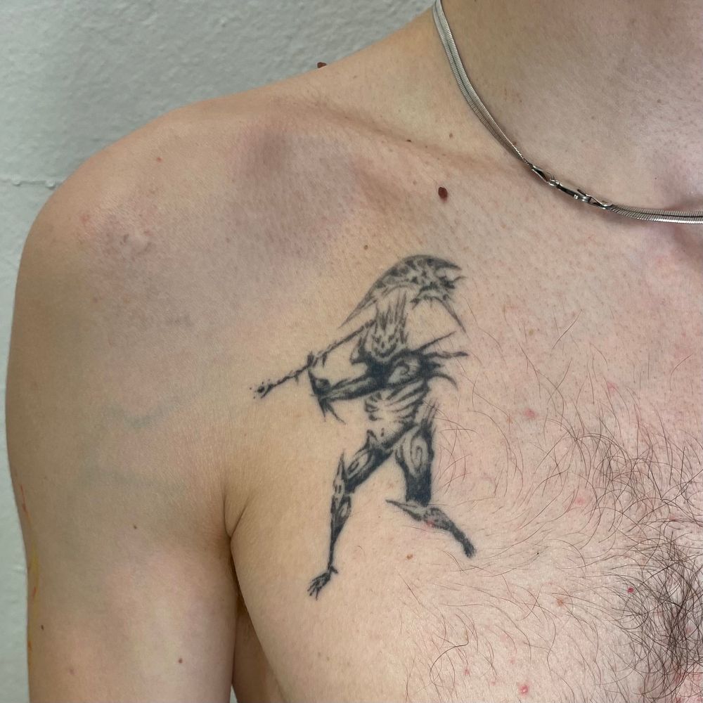 upper chest and shoulder with a healed black ink tattoo depicting a figure swinging a two handed axe. the figures wings has a skeletal face and is adorned with spikes and swirls. the figures wings is rendered with textural black marks and pepper shading. 