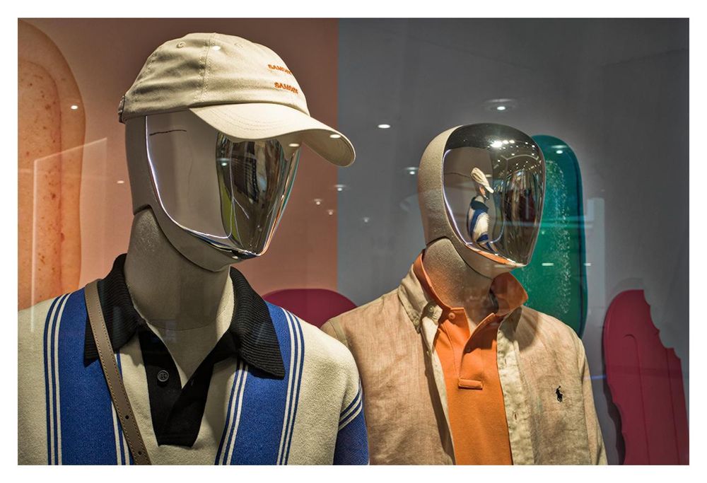 The image shows two mannequins dressed in stylish outfits, displayed in what appears to be a store window. The mannequin on the left is wearing a light beige cap, along with a clear, futuristic shiny chrome mask. Its outfit consists of a cream-colored polo shirt with blue and white vertical stripes and a black collar. A brown shoulder strap can be seen running above his shoulder. The mannequin on the right is dressed in a light beige jacket over an orange polo shirt. It also wears a shiny chrome mask that mirrors the surroundings, including a reflection of the left mannequin.