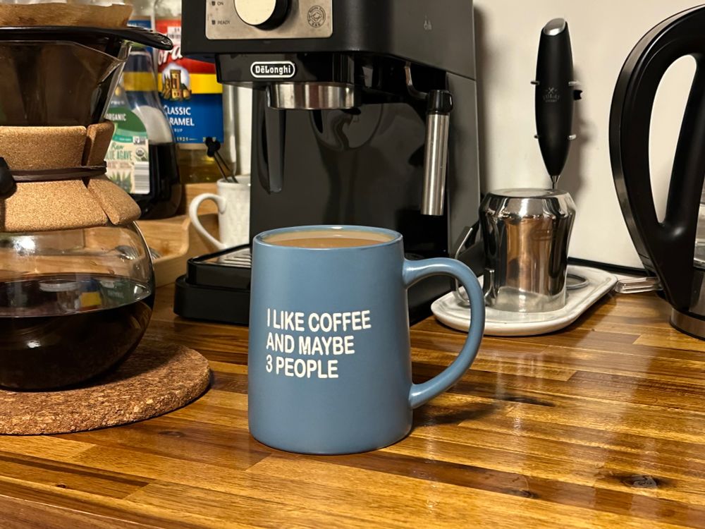 Blue coffe cup filled to the brim with coffee and cream, that has the words ‘I like coffee and maybe 3 people’ embossed on the front in white letters, sitting on a butcher block counter with various coffee accoutrements in the background. 