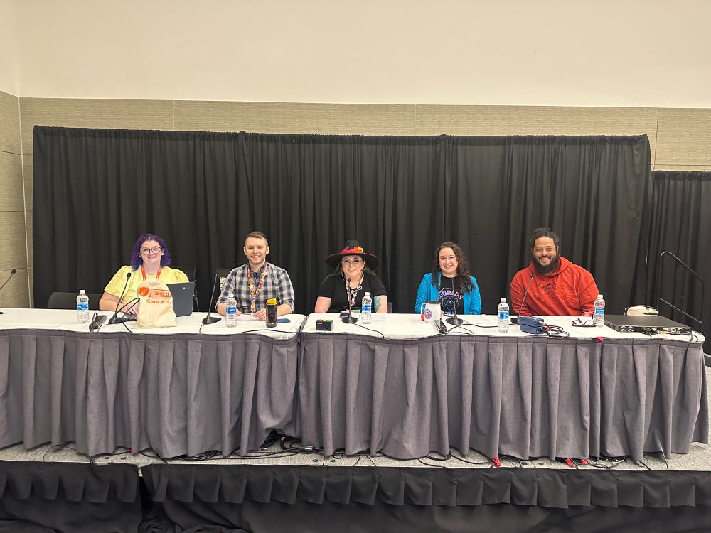 Five people sitting onstage at a long rectangular panel table smiling for the camera. 