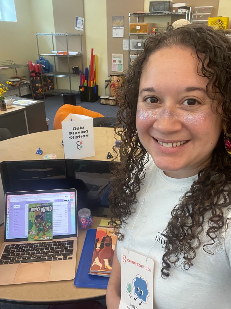 Selfie of a woman with brown curly hair, glitter makeup, and a white t-shirt posing next to a copy of the Nancy Druid TTRPG and a circular table with the sign “Role Playing Station”