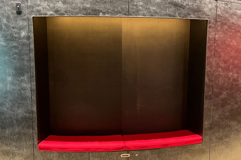 A colour photograph of a red cushioned seat within a recess in a wall. The lighting and textures of the recess and wall make the materials appear as though they’re made from metal 