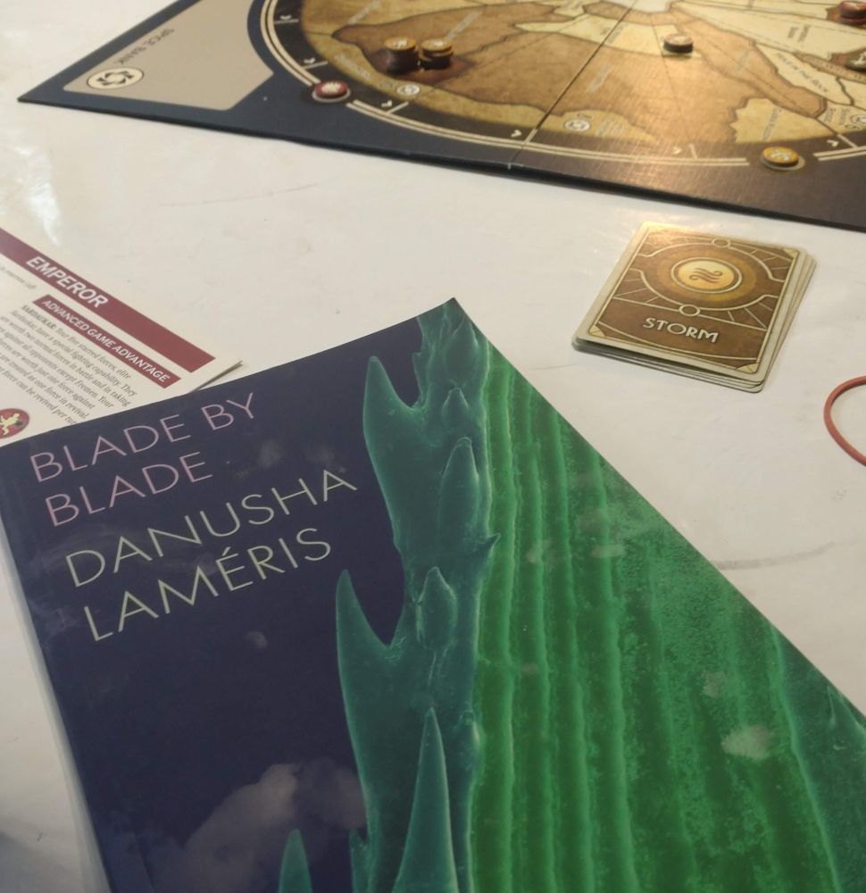 A copy of "Blade By Blade" by Danusha Laméris sitting on a table diagonally, in front of a board game on a white tablecloth. The board game is Dune. The book has a dark blue cover and against the blue background there is a magnified close up of a grass blade (or something similar) showing its tiny spurs.
