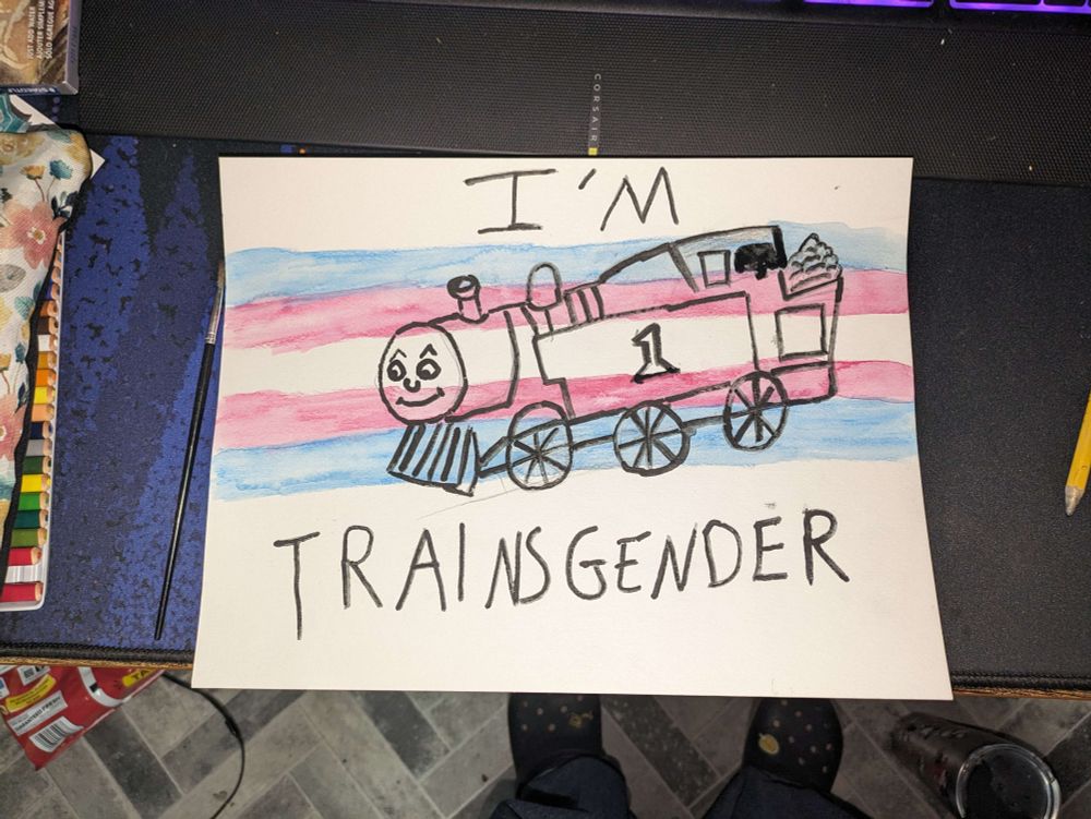 A water color painting of Thomas the Tank Engine with a trans flag over him. Text reads "I'M TRAINSGENDER"