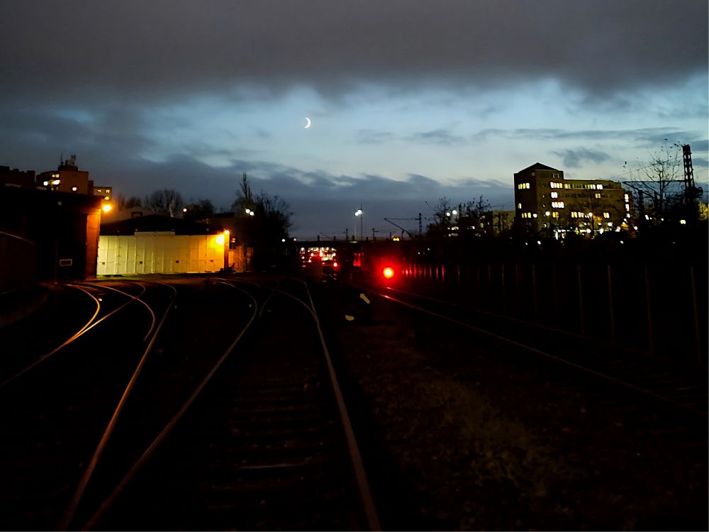 The image presents a nighttime scene of train tracks converging toward a distant red light, framed by buildings and illuminated by streetlights, under a partially cloudy sky.