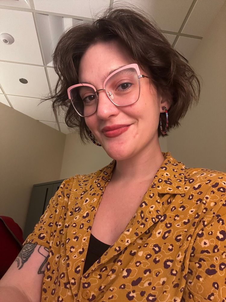 a selfie of me, a white lady with a short brown bob, wearing pink cat eye glasses, an orange and purple leopard print button up, big hoop earrings, and pink lipstick. i’ve got tattoos of a ribcage and a star trek badge barely visible on my right arm