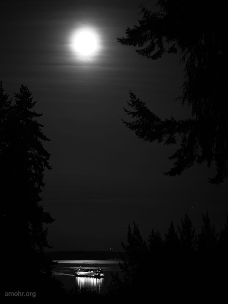 The last car ferry of the day passes under a bright glowy moon, streaked by thin hazy clouds. The ferry lights cast long bright reflections on a calm Puget Sound. Tall conifer trees reach their silhouettes into frame from left and right.