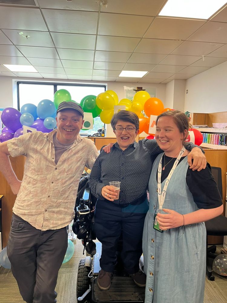 Amy stands in her electric wheelchair with the other members of the Pickering Lab. Amy has her arm around Niamh, a fellow PhD student. They are all smiling in front of a balloon arch that has a sign on it saying "Dr. Amy".