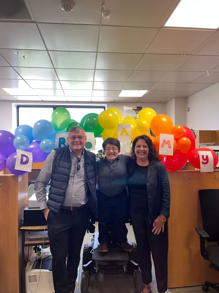 Amy stands in her electric wheelchair with her mam and dad standing either side of her. They are all smiling in front of a balloon arch that has a sign on it saying "Dr. Amy".