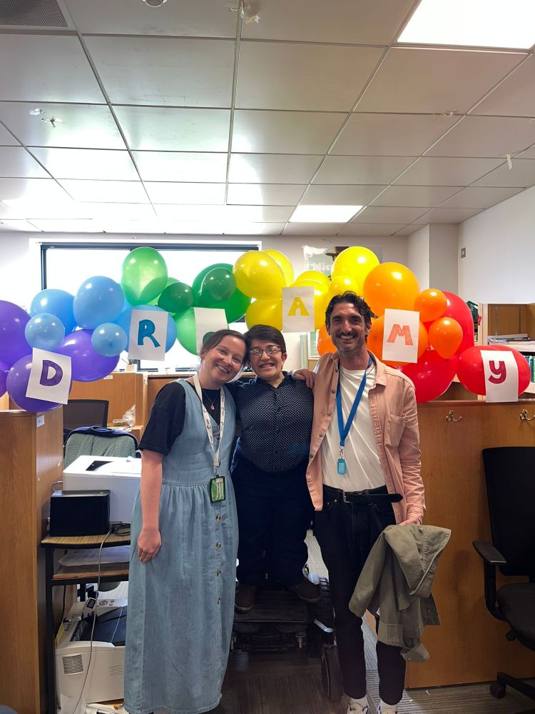 Amy stands in her electric wheelchair with the her arms around two friends, a man and a woman (Niamh and Mattia). They are all smiling in front of a balloon arch that has a sign on it saying "Dr. Amy".