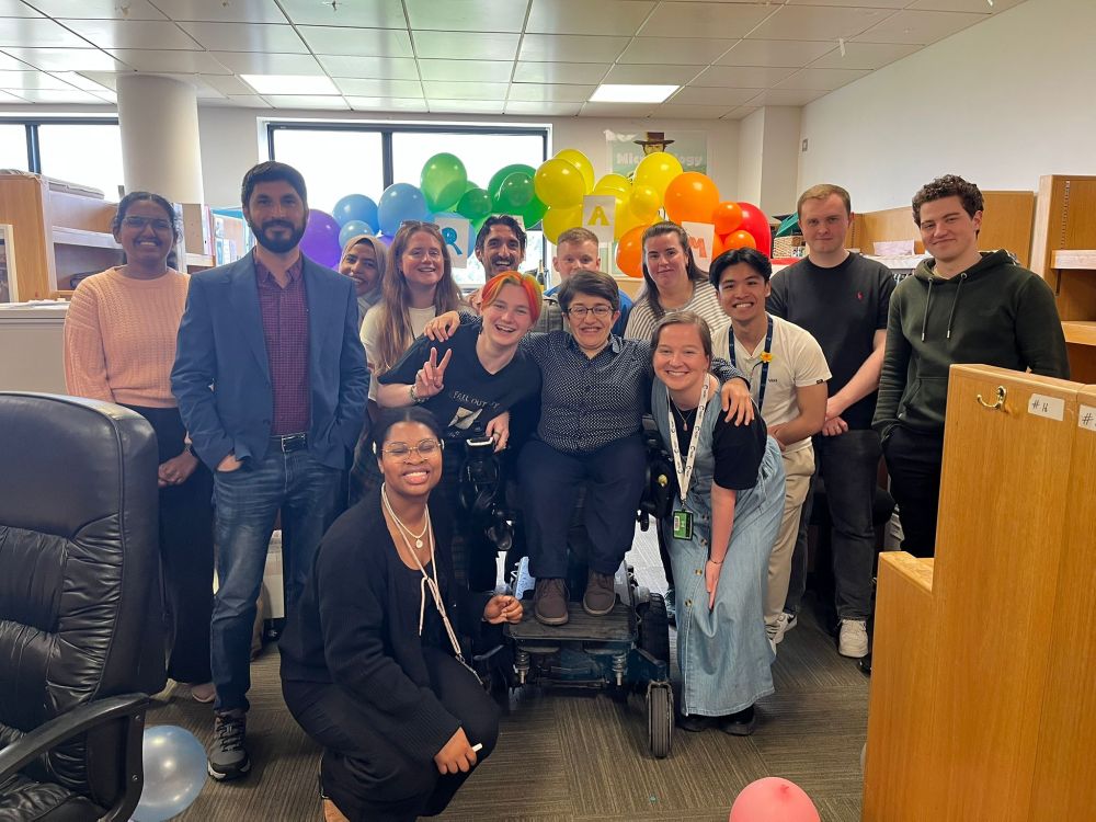Amy stands in her power wheelchair surrounded by a group of PhD students with her arms around two friends. They are all smiling in front of a balloon arch that has a sign on it saying "Dr. Amy".