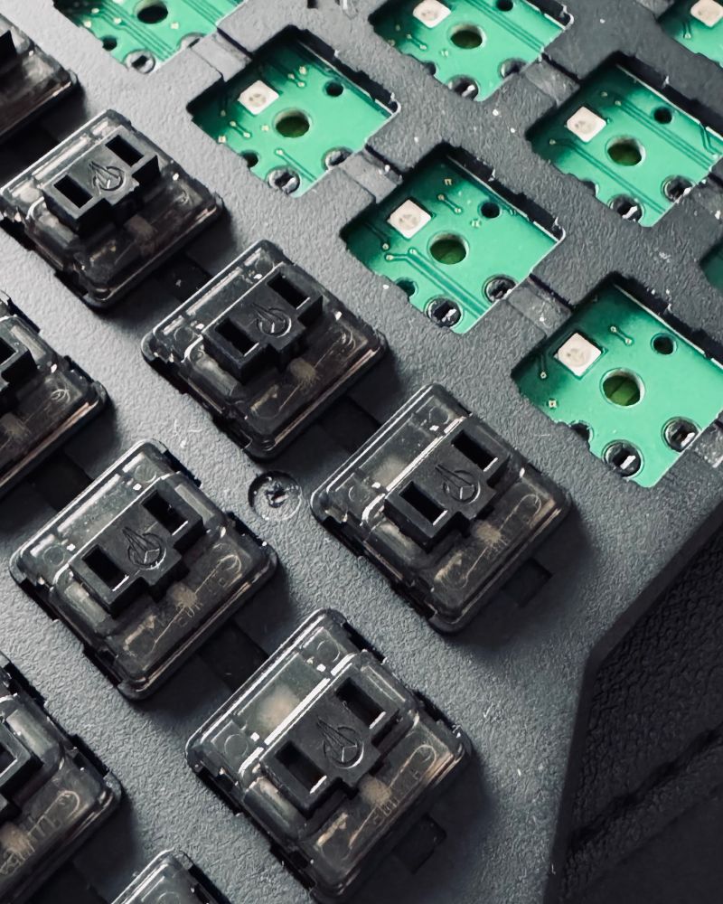 Close-up image of a mechanical keyboard's key switches and circuit board. The black key switches are arranged in a grid pattern, with some empty slots revealing green circuit board components underneath.