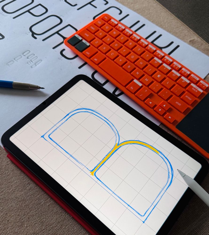 A tablet displaying a digital sketch of a letter “B” in Notes app, with a stylus resting on the screen. A printed type specimen sheet and an orange keyboard are in the background.