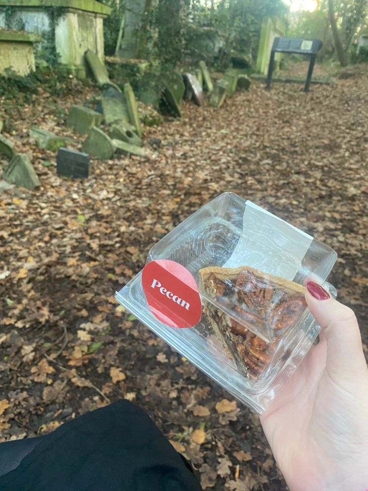 A white hand holding a clear box with a slice of pecan tart in it with a large circular red label with ‘Pecan’ written in white. 
There are leaves all over the path and grave stones and trees. A black sign in the distance.