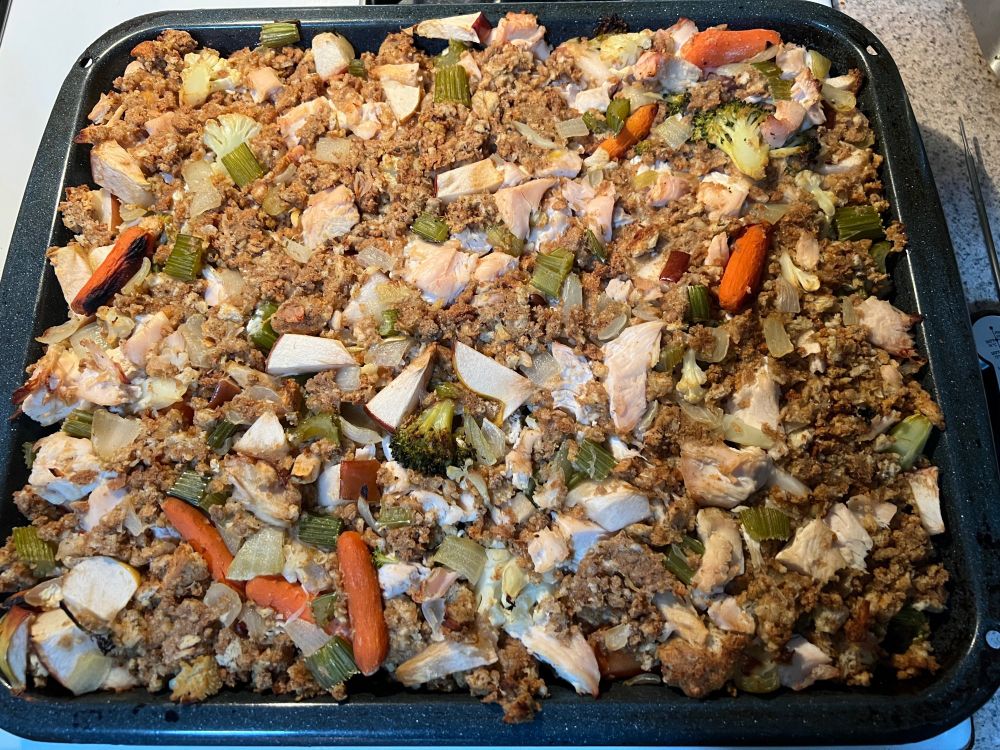 A broiler drip pan full of packaged bread stuffing augmented with chopped chicken breast, onion, celery, broccoli, cauliflower, and apple, whole baby-form carrots, and turkey gravy.