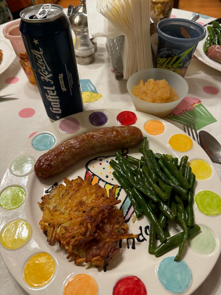 A rainbow polkadotted plate with a bratwurst sausage cooked green beans, and two misshapen potato pancakes. A cup of applesauce, and a German beer are in the background. All are on a cupcake tablecloth.