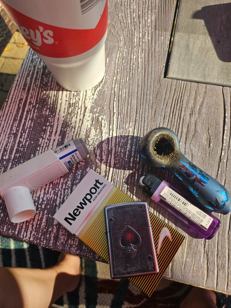 From upper left to bottom right, a Casey's medium sized drink, an albuterol inhaler, gold newport 100s with an ace of spades lighter on top, a purple bic lighter, and a cannabis pipe, empty but well used.

All of this is sitting on a grey and white wood patterned table with the legs of the photographer barely in frame at the bottom of the image