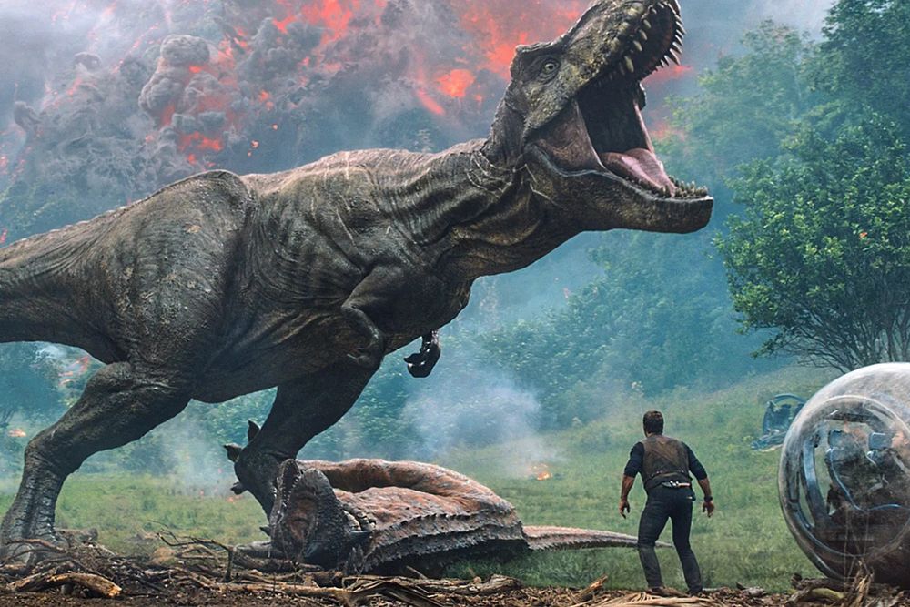 A large T-Rex steps on a Carnotaurus while Owen Grady (played by Chris Pratt) looks on. In the background is the beginning of a pyroclastic flow forming from an erupting volcano unseen. on the far right is a Gyrosphere holding two other characters