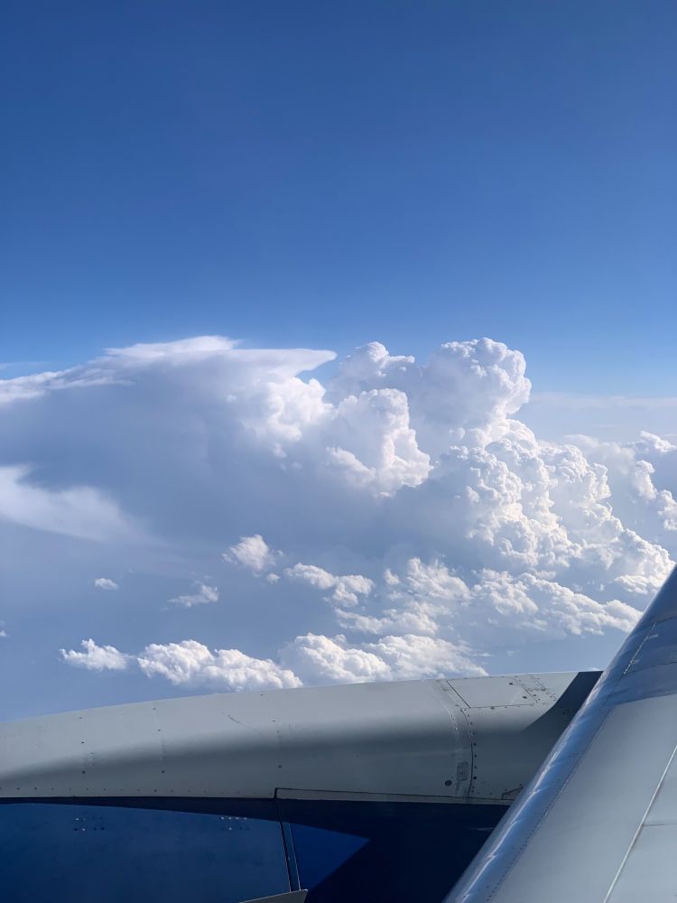 tall white fluffy cumulous clouds below a clear blue sky, as viewed from my airplane seat en route to Atlanta from LA; the top of a blue and silver plane engine and the edge of a silver wing fill the foreground