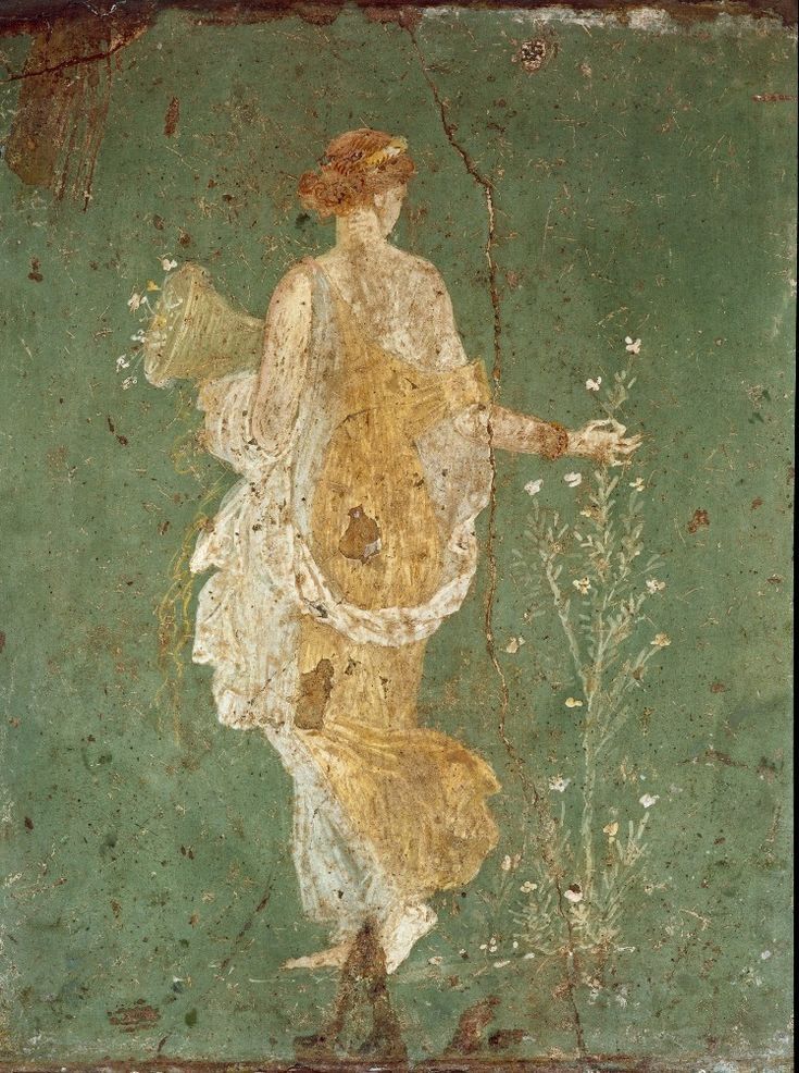 The back of Ostara in a Grecian style dress. Her face is turned away from the viewer and she is holding a container filled with flowers in one hand while the other reaches out to pick some flowers growing.