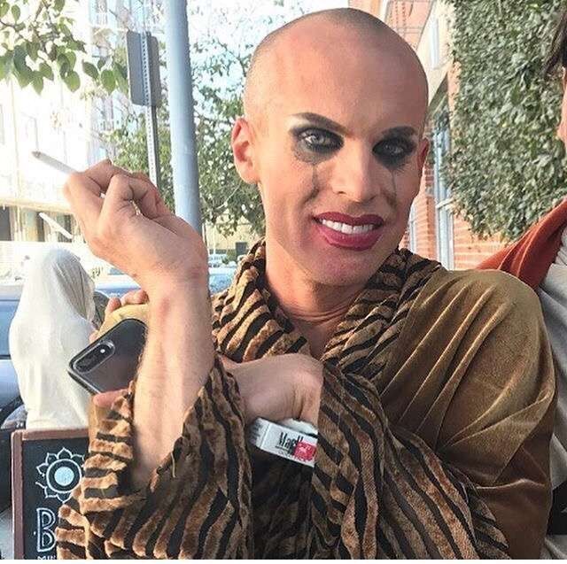 Drag queen Katya in a robe holding her phone and a pack of cigarettes in one hand. In the other is a lit cigarette. She has no wig on and her makeup is streaked down her face from crying but she is forcing a smile.