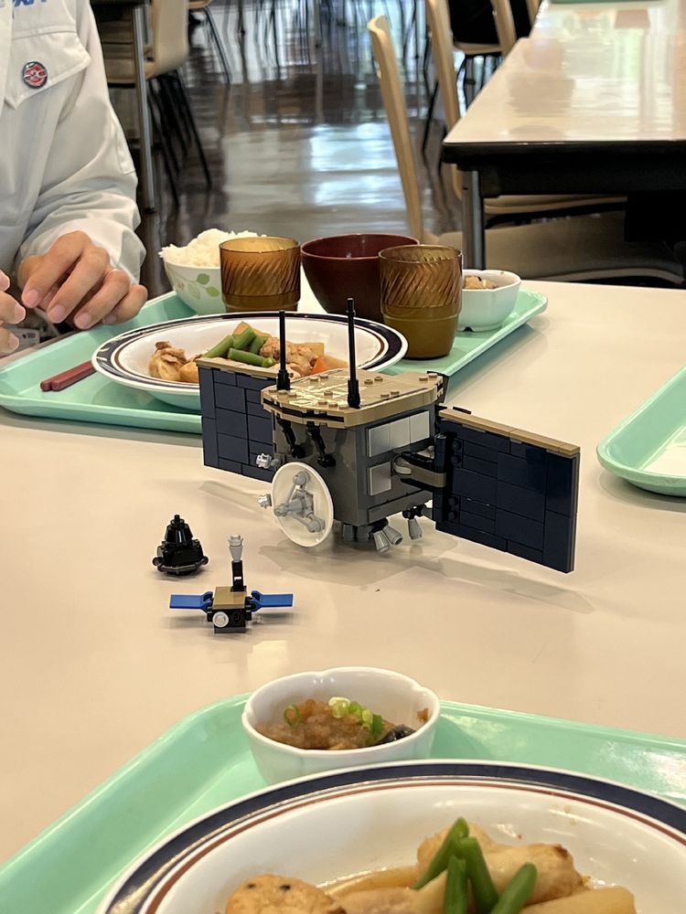Photograph showing a canteen table with the corners of three light green dinner trays showing. In the centre is a Lego model of a spacecraft with two small probes sitting beside it. 
