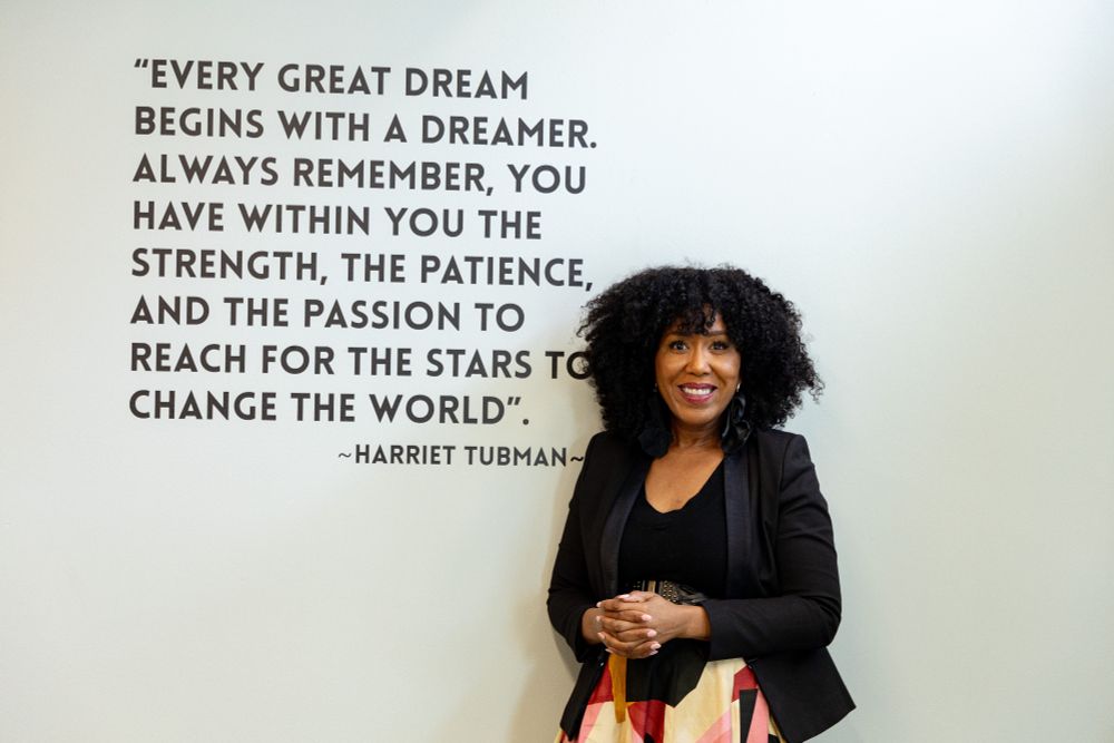 A picture of Dominique Samari standing in front of a wall with text that reads, "Every great dream begins with a dreamer. Always remember, you have within you the strength, the patience, and the passion to reach for the stars to change the world. - Harriot Tubman"