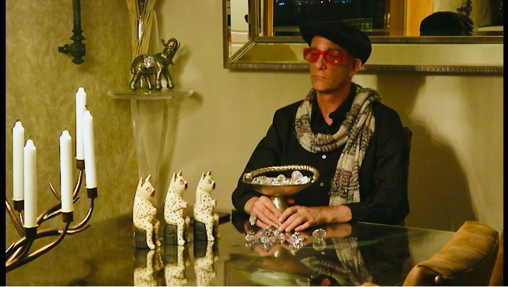 Man in a black beret, red sunglasses, and a scarf is sitting on a dining table holding a comically large goblet full of clearly fake jewels. There’s a statue of three… possibly hyenas(?) next to him