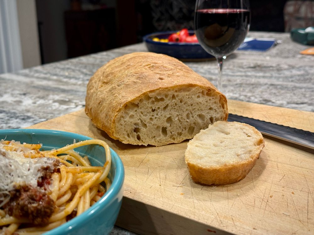 Oblique color photo of a kitchen counter still-life centering a sliced-open loaf of crusty  bread with an open and chewy crumb on a wooden cutting board, with a bowl of pasta in the lower left corner and a glass of red wine and shallow bowl of fresh tomatoes on a gneiss countertop behind the bread. 