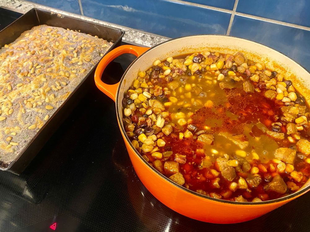 Color photo of a bright orange Dutch oven filled to the brim with posole in a red chile broth next to a gray steel rectangular pan of blue cornbread topped with melted cheddar cheese and toasted piñon, on a shiny black induction cooktop with a deep blue tile backsplash and thin strip of polydeformed migmatitic biotite gneiss. 
