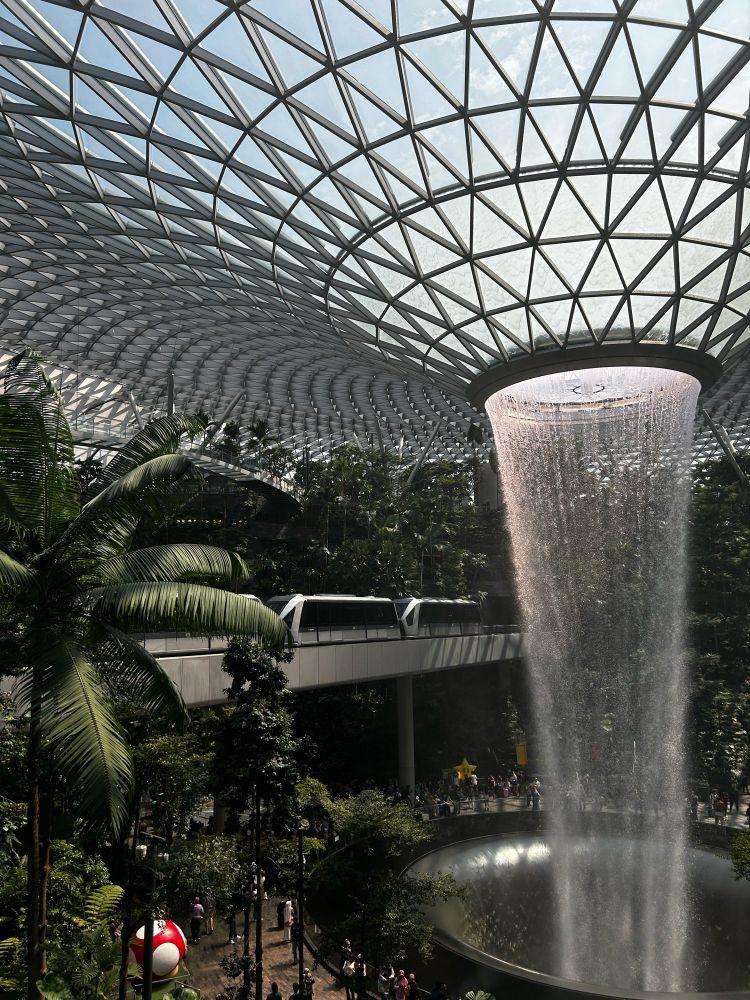Photo of the waterfall and train in Changi Airport in Singapore. You can see visitors walking around through trees and there is a Mario theme going on. 