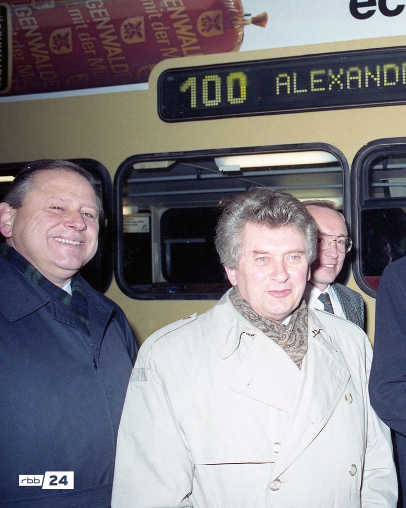 Mehrere Männer in Jacken und Mänteln stehen lächelnd vor einem gelben Doppeldeckerbus der Berliner Buslinie 100. Auf dem digitalen Zielanzeiger des Busses steht „100 Alexanderplatz“. Im Hintergrund ist ein Werbeplakat mit einer Wurst und dem Schriftzug „GENWALD“ zu sehen. Unten links ist das Logo von rbb24 eingeblendet. Die Aufnahme wirkt wie ein historisches Pressefoto, vermutlich zur Eröffnung oder einem Jubiläum der Buslinie.