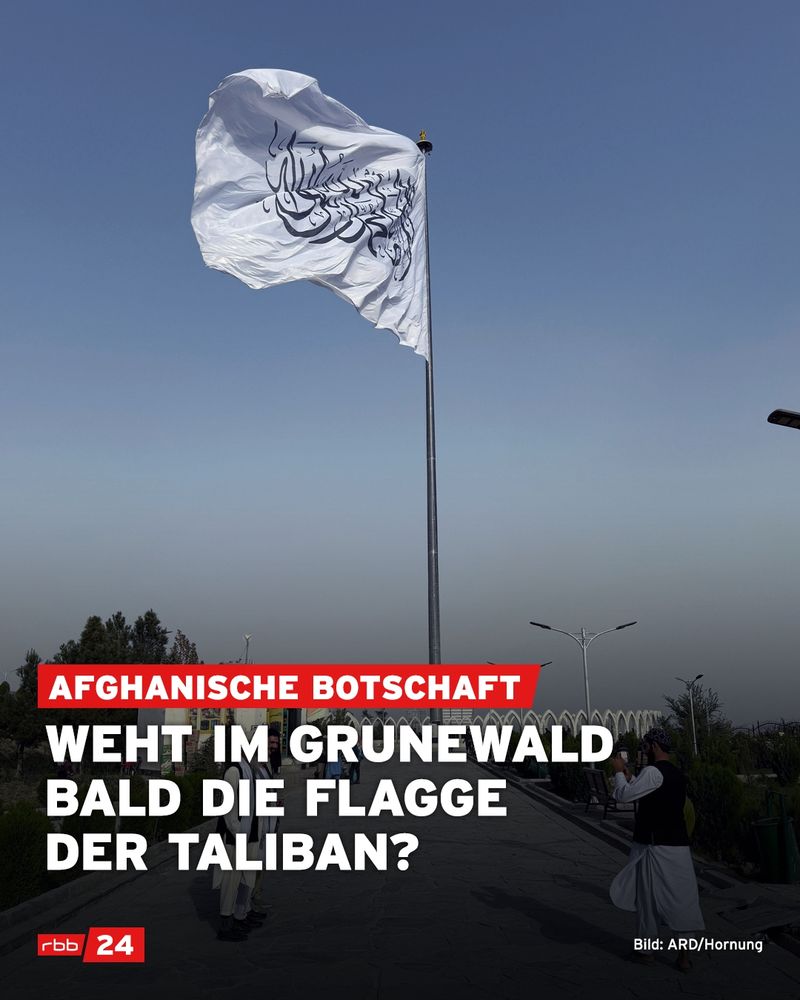 Große weiße Flagge mit schwarzer arabischer Schrift, Symbol der Taliban, weht an einem hohen Mast vor blauem Himmel. Im Vordergrund ein Gehweg mit mehreren Personen in traditioneller afghanischer Kleidung. Im unteren Bildteil steht in weißer und roter Schrift auf schwarzem Hintergrund: „Afghanische Botschaft – Weht im Grunewald bald die Flagge der Taliban?“ Links unten das Logo von „rbb24“, rechts unten die Bildquelle: „Bild: ARD/Hornung“.